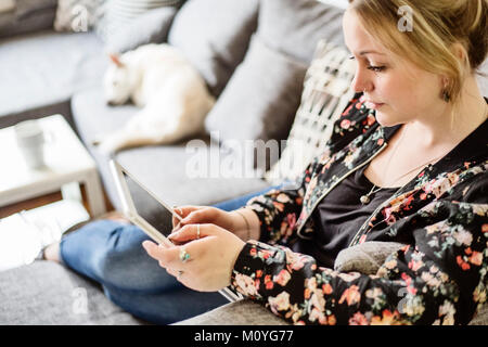 Junge Frau sitzt auf dem Sofa, mit ihrem Hund und schaut auf einen Tablet-PC, Deutschland Stockfoto