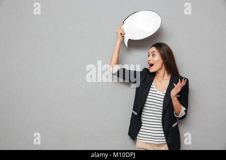 Asian Business woman holding leere Sprechblase Overhead und weg schauen entsetzt über grauer Hintergrund Stockfoto