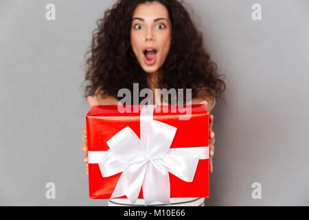 Brünette Frau in Defokussieren gegen graue Wand holding red box Geschenk - Posing mit weißen Bogen gewickelt, es mit der Kamera Stockfoto