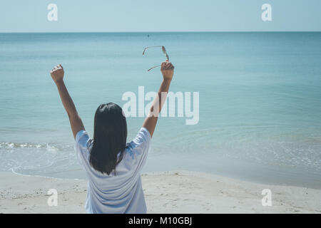 Und Urlaub Konzept: Frau tragen weiße T-Shirt, Sie auf Sand Strand und Holding Sonnenbrille in ihrer Hand, sie mit Blick auf das Meer und Stockfoto