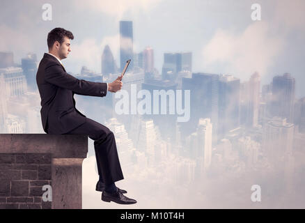 Ein junger Geschäftsmann am Rande eines Gebäudes vor einem Stadtzentrum scape Hintergrund mit hohen Gebäuden und Wolken Konzept sitzen Stockfoto