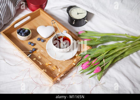 Ein Tablett mit Kaffee und Lebkuchen in der Form von Herzen und einem Blumenstrauß aus Tulpen auf dem Bett Stockfoto