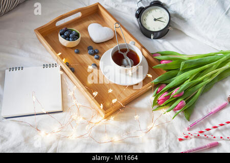 Ein Tablett mit Kaffee und Lebkuchen in der Form von Herzen und einem Blumenstrauß aus Tulpen auf dem Bett Stockfoto