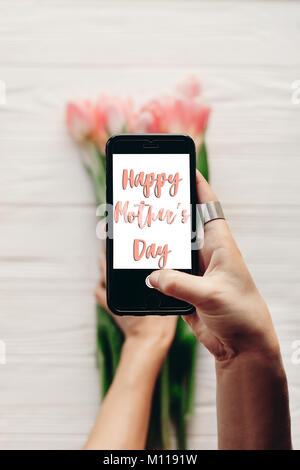 Happy Mother's Day text Zeichen auf dem Bildschirm des Telefons. Hand Gadget mit Meldung über rosa Tulpen auf weißen Holzmöbeln rustikal Hintergrund. Grußkarte Stockfoto