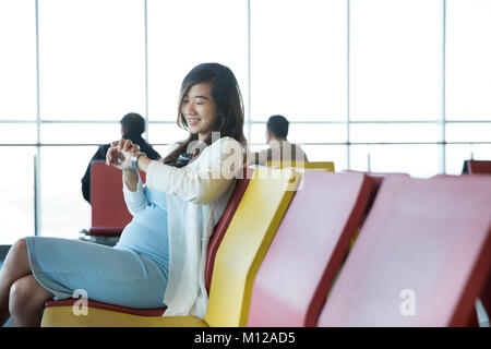 Frau im Flughafen Lounge im Stuhl sitzend durch das Fenster schaut auf ihre Uhr warten auf Ihren Zeitplan zu fliegen Stockfoto