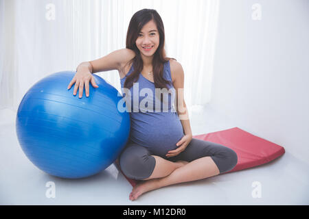 Das Portrait einer schönen asiatischen schwangere Frau sitzt auf einem Yoga Matte, eine Kugel und lächelnd Stockfoto