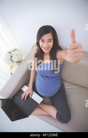 Das Portrait einer schönen schwanger asiatische Frau sitzt auf einem Sofa mit Laptop auf dem Schoß, Daumen hoch zufrieden Geste Stockfoto