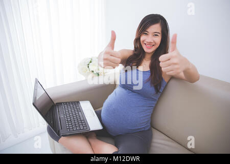 Das Portrait einer schönen schwanger asiatische Frau sitzt auf einem Sofa mit Laptop auf dem Schoß, Daumen hoch zufrieden Geste Stockfoto