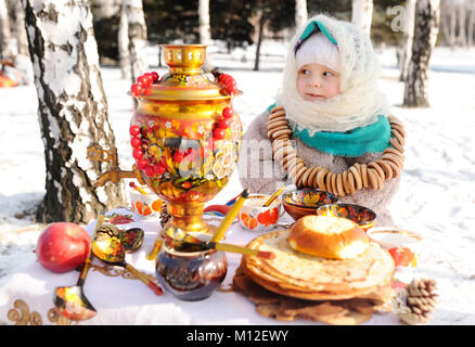 Kind, Mädchen in einem Pelzmantel und in einen Schal im russischen Stil mit einem großen Samovar in den Händen der Pfannkuchen mit roter Kaviar, matryoshkas und Geschirr in Khokhloma Stil Stockfoto