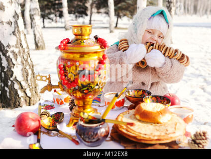 Kind, Mädchen in einem Pelzmantel und in einen Schal im russischen Stil mit einem großen Samovar in den Händen der Pfannkuchen mit roter Kaviar, matryoshkas und Geschirr in Khokhloma Stil Stockfoto