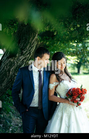 Sensible Outdoor Portrait des wunderschönen Brautpaar umarmen unter dem Baum. Der lächelnde brunette Braut hält und mit Blick auf die Hochzeit Blumenstrauß aus roten und rosa Blüten. Stockfoto