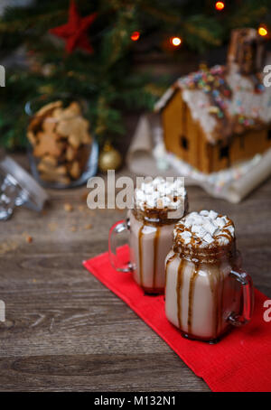 Zwei Gläser Kakao mit Marshmallows, Sahne und Schokolade Sirup auf Holztisch und Lebkuchenhaus, Cookie Jar und Weihnachtsbaum mit zu Stockfoto