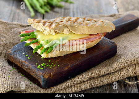 Street Food: heiße Panini mit Schinken und grünem Spargel überbacken mit geriebenem Käse auf einem hölzernen Hintergrund serviert. Stockfoto