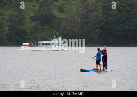 2 Frauen stehen auf paddleboards auf Coniston Water während Leute auf Launch Boot Kreuzfahrt Reise segeln Darüber hinaus - Lake District, Cumbria, England, UK. Stockfoto