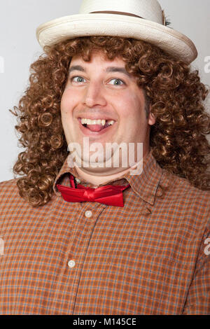 Mann, gekleidet in ein albernes Kostüm, mit einem karierten Hemd, Roter Fliege, lange lockige Haare, einen Strohhut und einem großen Lächeln. Stockfoto