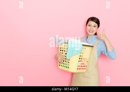 Sicher gerne weibliche Wäscheservice store Arbeiter holding Bekleidung Warenkorb an Kamera und zeigen "Daumen hoch" Geste auf rosa Hintergrund isoliert. Stockfoto