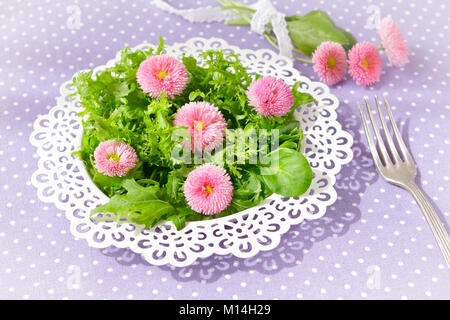Weiße Platte mit einem Salat von Gemischte grüne Salatblätter und genießbare daisy flowers, zusammen mit einer alten Gabel auf eine nostalgische lila Hintergrund Stockfoto
