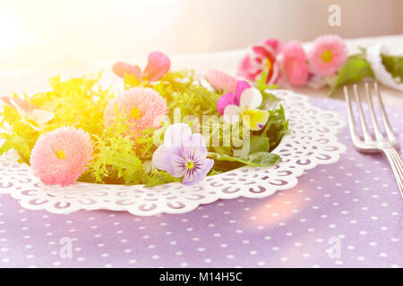 Weiße Platte mit einem Salat von Gemischte grüne Salatblätter und genießbare Gänseblümchen und Stiefmütterchen Blumen, zusammen mit einer alten Gabel auf eine nostalgische Lila backgroun Stockfoto