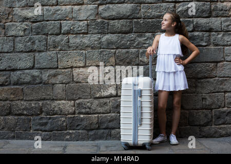 Gerne kleine Mädchen in Weiß mit Reisetasche auf Stein Wand Hintergrund Stockfoto