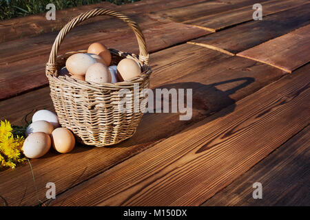 Frisches Huhn Eier im Korb auf rustikalen Holzmöbeln Hintergrund. Blumenstrauß mit gelben Löwenzahn Stockfoto