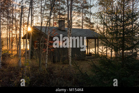 Finnische Landschaft: Traditionelle lakeside Blockhaussauna an einem abgeschiedenen Ort versteckt am Rande der Herbst/Winter durch die Bäume, in die Sonne, Finnland Stockfoto