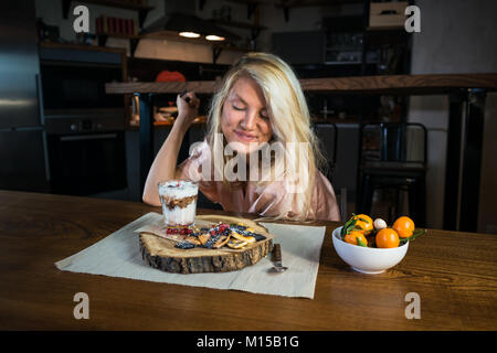 Junge Frau lächelt und isst Frühstück im Wohnzimmer zu Hause. Lustige Blondine sitzt am Tisch mit Essen gegen moderne Küche in den frühen Morgenstunden Stockfoto