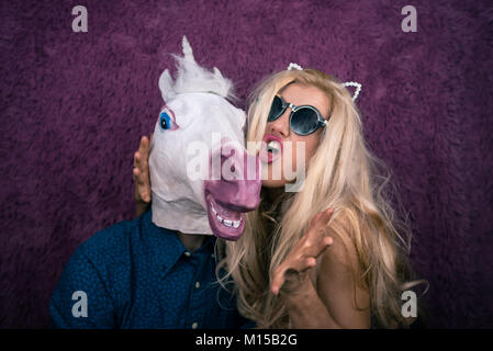 Portrait von Happy Einhorn in den Armen von freaky junge Frau auf dem violetten Hintergrund. Expressive Blondine mit seltsamen Kerl. Ungewöhnliche Menschen zeigt Emotionen Stockfoto