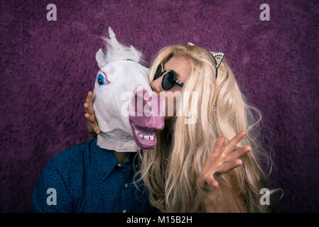 Freaky junge Frau küssen glücklich Einhorn auf dem violetten Hintergrund. Expressive Blondine mit seltsamen Kerl. Ungewöhnliche Menschen sitzt zusammen Stockfoto