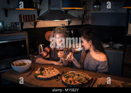 Zwei junge Frauen am Tisch mit Essen und Trinken Wein sitzen. Romantische blonde und brünette Dining am Abend. Wein zu Hause prüfen Stockfoto