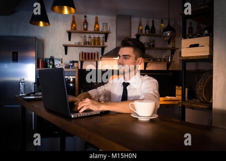 Junger Mann sitzt in der Küche, an der Theke und arbeitet in Laptop. Home Atmosphäre, Arbeit am Abend nach einem anstrengenden Tag bei einer Tasse Kaffee Stockfoto