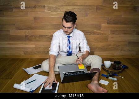 Junge Mann arbeitet in lustiger Kleidung. Eine barfuß Geschäftsmann, sitzen auf dem Boden an einer Wand und der Erstellung eines Laptop Stockfoto