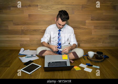 Eine barfuß Geschäftsmann sitzt auf dem Boden an einer Wand mit Laptop und anderen Gadgets und schneidet ein Blatt. Ernsthafte junge Mann nach der Arbeit in den lustigen Kleidung Stockfoto