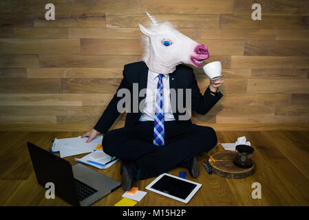 Junger Mann, der lustige Maske sitzt auf dem Boden gegen eine Wand und trinkt Kaffee. Unicorn in Anzug und Krawatte arbeiten im Home Office Stockfoto
