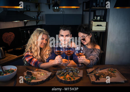 Zwei Frauen mit einem Mann lächelt und hat ein gemeinsames Abendessen. Gruppe von glücklichen Menschen in die Kamera schauen und hält Gläser Wein. Die jungen Menschen Zeit verbringen Stockfoto