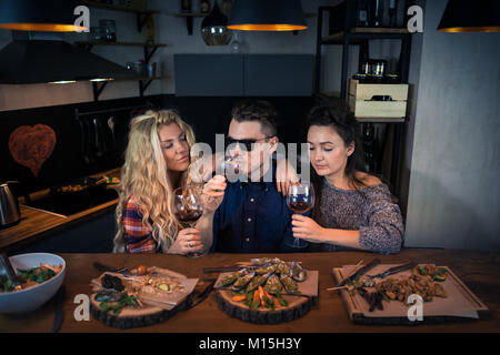 Zwei junge Frauen mit einem Mann in Sonnenbrille hat spät Abendessen in Buffetform in einer modernen Küche zu Hause. Gruppe von schönen Menschen sitzt zusammen und isst Essen mit Wein Stockfoto