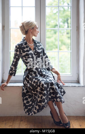 Schöne blonde Frau in Kleid sitzt am Fenster Stockfoto