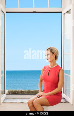 Blonde Frau in einem roten Kleid sitzt am offenen Fenster mit Meerblick Stockfoto