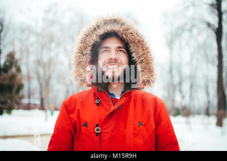 Porträt, Nahaufnahme der Jungen stilvoll Mann lächelnd mit einem Bart rot Winter Jacke mit Kapuze und Fell auf dem Kopf bekleidet steht gegen die backgro Stockfoto