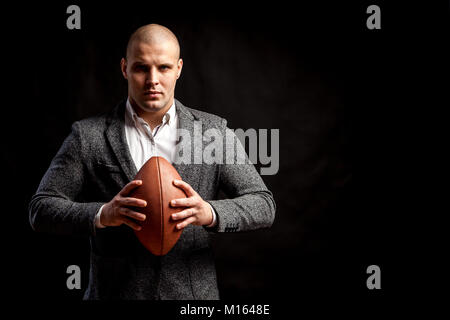 Eine junge glatzköpfige Mann in einem weißen Hemd, grauen Anzug hält einen Rugby Ball auf einem schwarzen Hintergrund isoliert Stockfoto