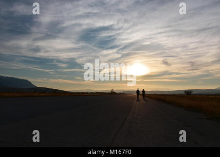 Sonnenuntergang, flachen Tal, zwei Menschen zu Fuß auf eine breite, gerade, leere asphaltierte Straße in Richtung der Sonne am Horizont Stockfoto