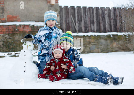 Kleinen niedlichen Lächeln baby boy und seine beiden älteren Brüder, sitzen draußen im Schnee, Schneemann neben Ihnen Stockfoto