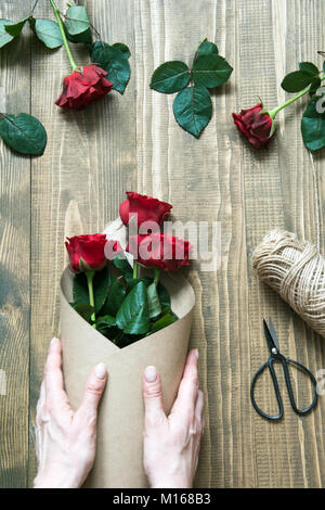 Florist eine rote Rosen Blumenstrauß, Umwickeln mit Kraftpapier auf einem Holztisch. Blick von oben. Im rustikalen Stil. Stockfoto