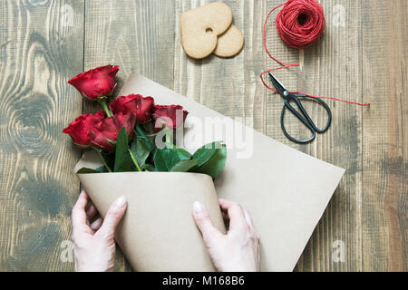 Florist eine rote Rosen Blumenstrauß, Umwickeln mit Kraftpapier auf einem Holztisch. Blick von oben. Im rustikalen Stil. Stockfoto