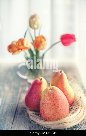 Eine Gruppe von schönen reife, gelbe Birne lag auf einem hölzernen Tisch in einen beleuchteten Zimmer im rustikalen Stil auf dem Hintergrund einer schönen Blumenstrauß orange Tulpen. Kopieren Sie Platz. Stockfoto