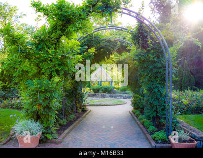 Sonnenuntergang scheint durch einen Torbogen. Einen Stein Weg führt zu einem schönen Garten, der sich um einen Pavillon zentriert ist. Stockfoto