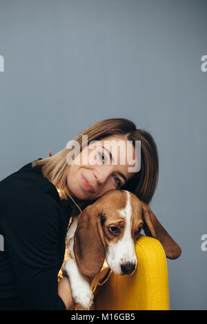 Junge Frau in schwarzem Kleid Umarmungen Beagle Welpen auf gelb Stuhl mit grauen Wand im Hintergrund Stockfoto