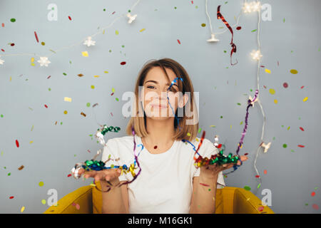 Junge Frau im weißen T-Shirt blast Konfetti auf gelb Stuhl mit grauen Wand im Hintergrund Stockfoto