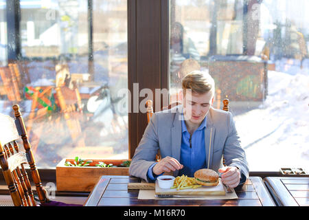 Portrait von busy Young Business Mann sitzend Mit einem Glas trinken und Hamburger im Fast Food Restaurant. Attraktive kaukasischen Mann essen und mit Notebook in ein Cafe. Im Innenbereich Stockfoto