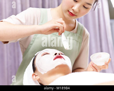 Asiatische Kosmetikerin Anwendung weiße Gesichtsmaske mit einem Pinsel auf Gesicht einer jungen Frau. Stockfoto