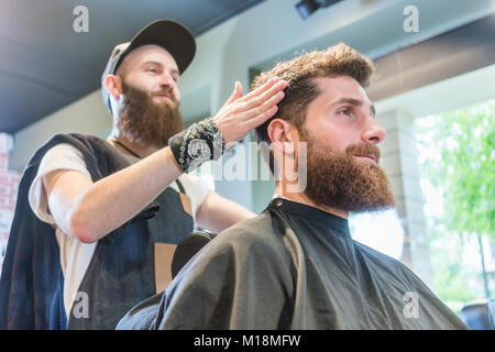 Gut aussehender bärtiger junger Mann bereit für einen modischen Haarschnitt in einem kühlen Stockfoto